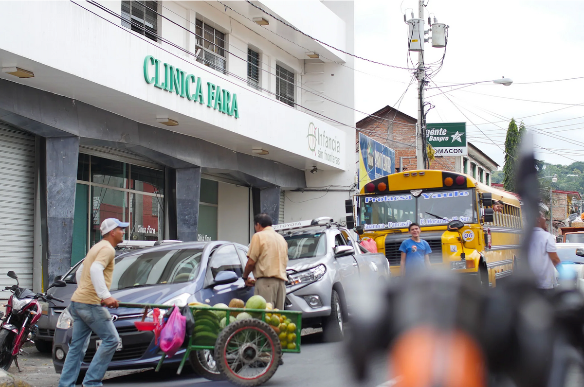 Busy main street in a town at origin, with a fruit cart, parked cars and a local bus